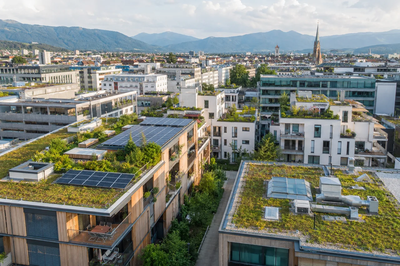 Luftaufnahme eines dicht bebauten Stadtquartiers, in dem zahlreiche Wohn- und Bürogebäude städtische Dachbegrünungen und Solaranlagen auf ihren Dächern tragen, umrahmt von Bergen am Horizont.