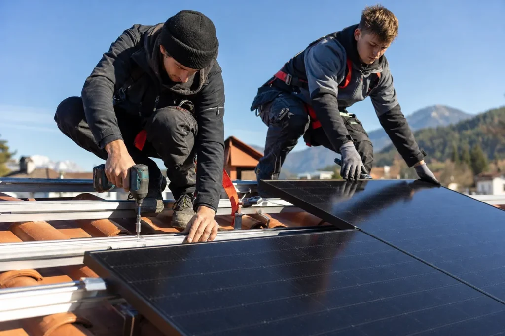 Zwei Handwerker montieren bei der Auf-Dach-Photovoltaik schwarze PV-Module auf den Montageschienen eines Ziegeldachs, im Hintergrund die Berge.