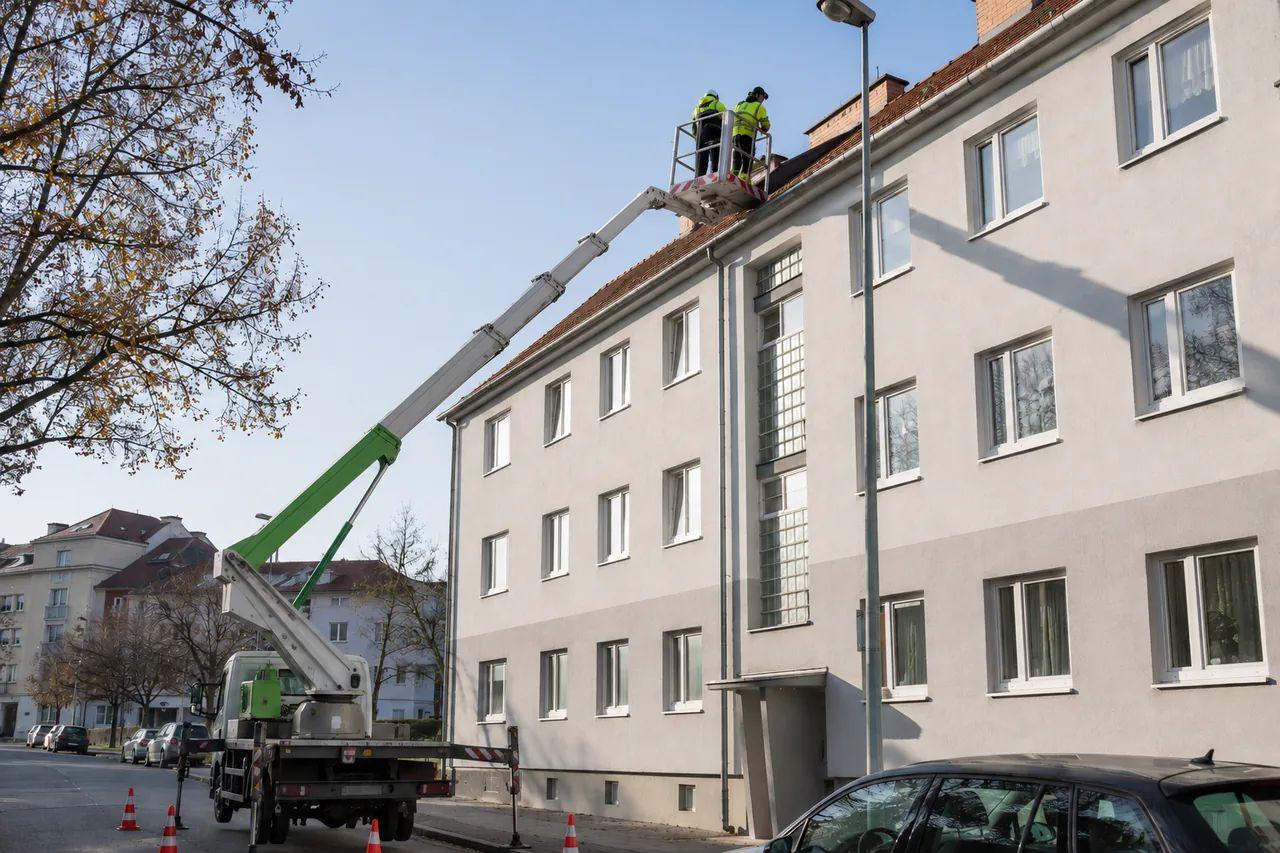 Zwei Arbeiter in Warnwesten reinigen von einer Hubarbeitsbühne aus die Dachrinne eines mehrstöckigen Mietshauses und zeigen die professionelle Dachrinnenreinigung im Mehrparteienhaus.