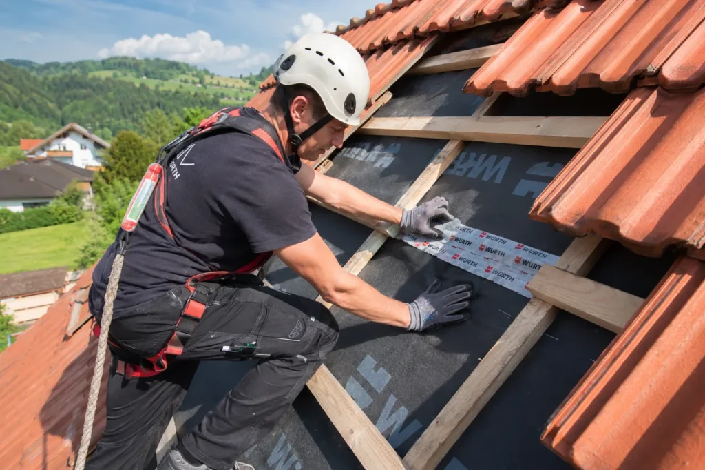 Ein Dachdecker mit Helm und Auffanggurt legt für eine Dachreparatur einige rote Ziegel zurück und dichtet die freigelegte Unterdeckbahn an der schadhaften Stelle ab.