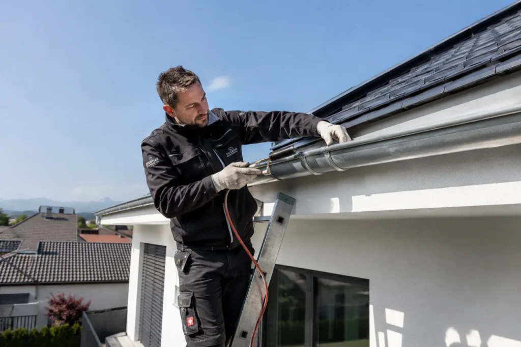 Ein Spengler arbeitet von der Leiter aus an der Metall-Dachrinne und der Traufverblechung eines Hauses und dichtet den Anschluss fachgerecht ab.