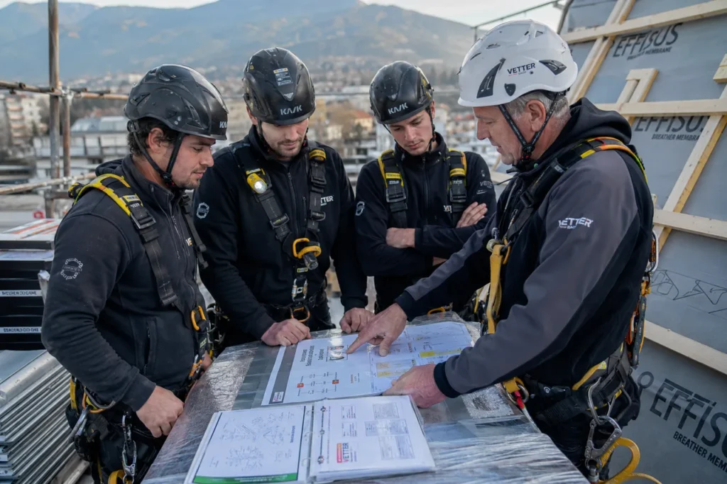 Vier Dachdecker in Schutzhelmen und Auffanggurten besprechen bei einer Sicherheitsunterweisung auf dem Dach gemeinsam einen ausgebreiteten Montageplan zur Arbeitssicherheit.