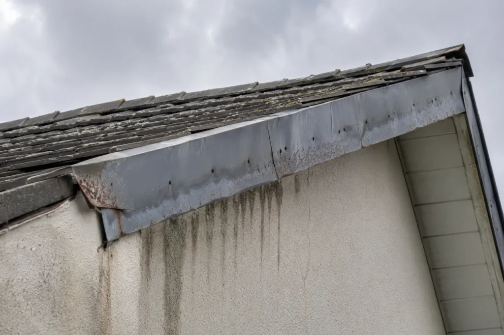 Altes, verrostetes und verformtes Ortgangblech an einer Dachkante, unter dem Feuchtigkeit als dunkle Wasserspuren die Giebelwand hinabläuft.